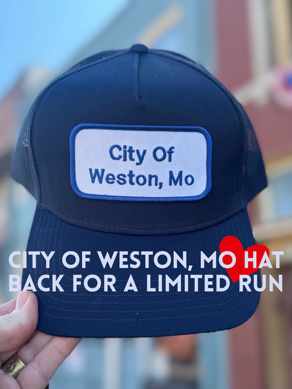 Navy blue cap with 'City of Weston, Mo' patch held by a hand against a blurred indoor background.