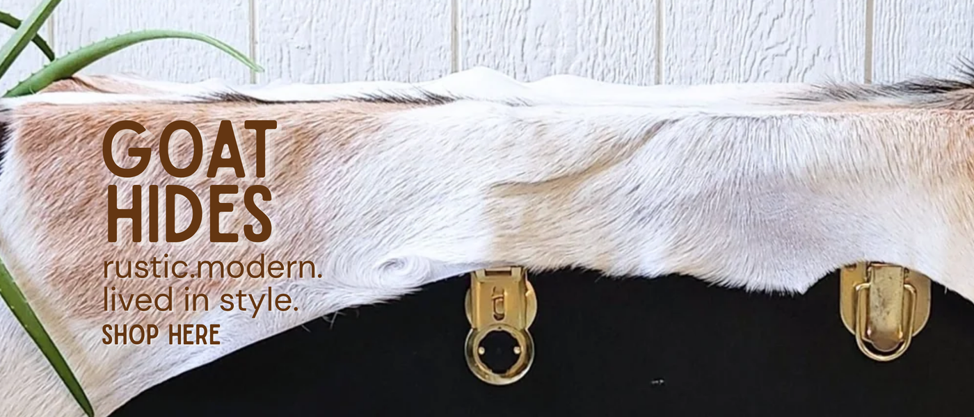 A large goat hide rug or wall covering in white, beige, and brown tones, draped across a black surface with two gold metal rings below. Text on the left reads 'GOAT HIDES rustic modern.